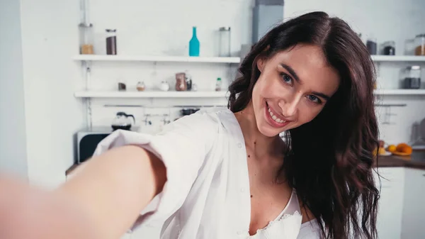 Brunette woman in white bra and unbuttoned shirt taking selfie while looking at camera on blurred foreground — Stock Photo