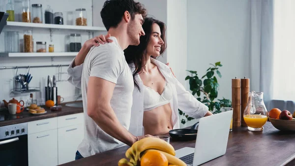 Mujer feliz en camisa blanca abrazando hombro de hombre cerca de la computadora portátil y frutas en la cocina - foto de stock