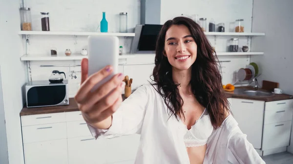 Mujer alegre en sujetador y camisa blanca tomando selfie en el teléfono celular en la cocina - foto de stock