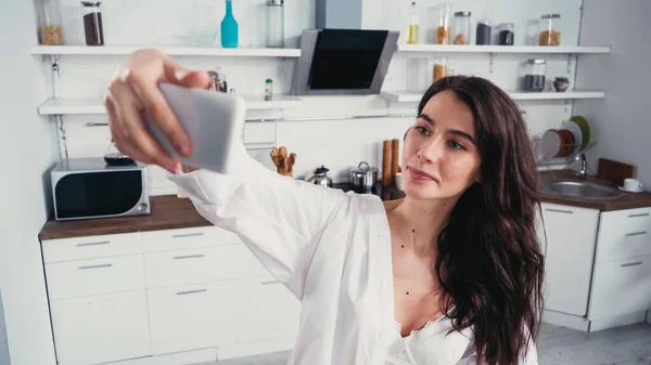Morena mujer en camisa blanca y sujetador tomando selfie en teléfono inteligente en la cocina - foto de stock