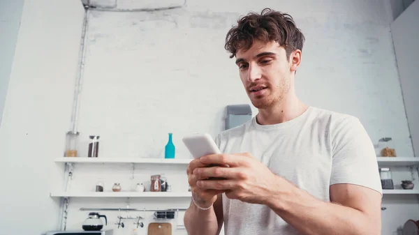 Vista de ángulo bajo de hombre joven en la camiseta blanca de mensajería en el teléfono inteligente en la cocina - foto de stock