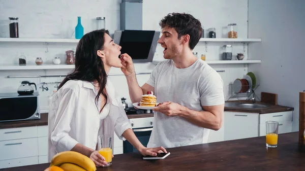 Young man with pancakes feeding sexy girlfriend with raspberry in kitchen — Stock Photo