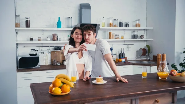 Donna sorridente in camicia e reggiseno scattare selfie con fidanzato vicino frittelle e frutta — Foto stock