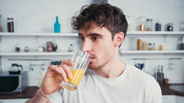 Young man in white t-shirt drinking fresh orange juice in kitchen — Stock Photo