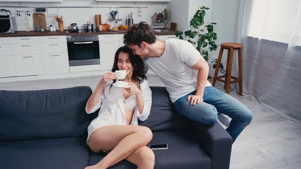Homem feliz perto mulher satisfeita beber café no sofá em camisa branca e sutiã — Fotografia de Stock