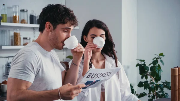 Joven pareja leyendo el periódico mientras bebe café de la mañana en la cocina - foto de stock