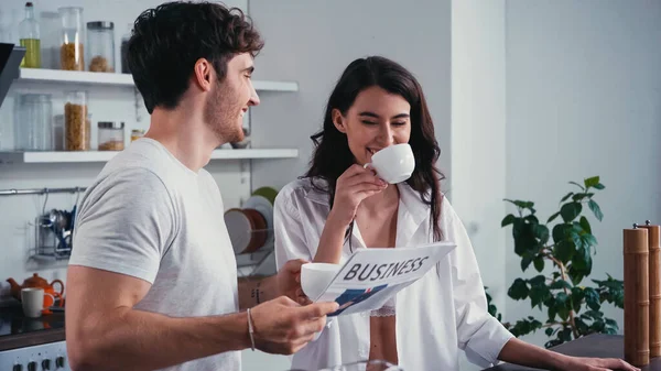 Улыбающийся мужчина держит деловую газету возле счастливой женщины пьет утренний кофе — стоковое фото