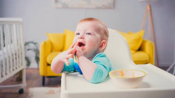 Menino infantil espantado com olhos azuis e boca aberta sentado na cadeira de alimentação com colher perto da tigela — Fotografia de Stock