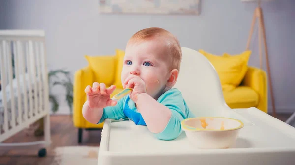 Menino com olhos azuis sentado na cadeira de alimentação e chupando colher perto tigela com comida de bebê — Fotografia de Stock