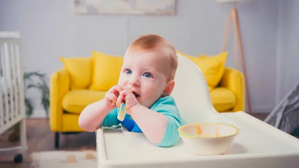Menino com olhos azuis sentado na cadeira de alimentação e chupando colher perto da tigela — Fotografia de Stock