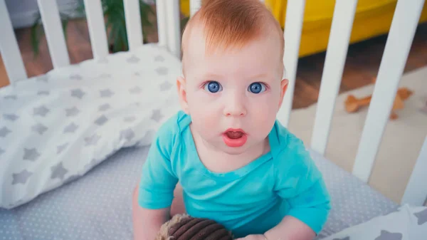 Высокий угол обзора удивленного мальчика, смотрящего в камеру и играющего с мягкой игрушкой в размытой кроватке — стоковое фото