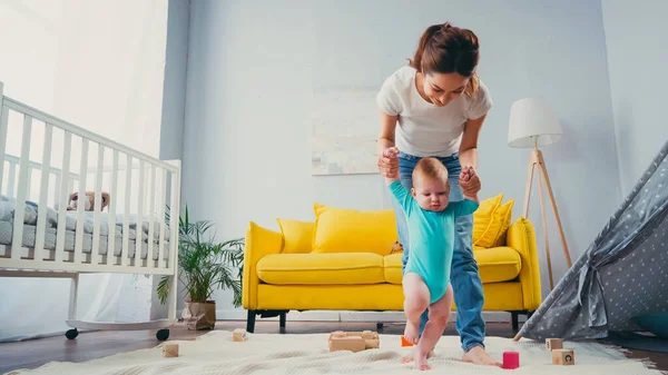 Felice madre che si tiene per mano e sostiene il figlio neonato che cammina sulla coperta vicino ai giocattoli — Foto stock