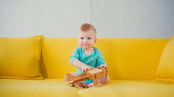 Niño feliz niño sentado en el sofá y jugando con biplano de madera - foto de stock