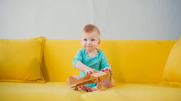 Happy infant boy sitting on sofa and playing with wooden biplane — Stock Photo