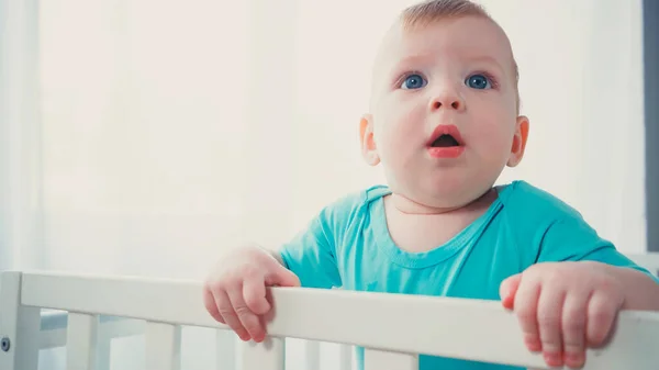 Sorprendido bebé niño de pie en la cuna del bebé y mirando hacia arriba - foto de stock