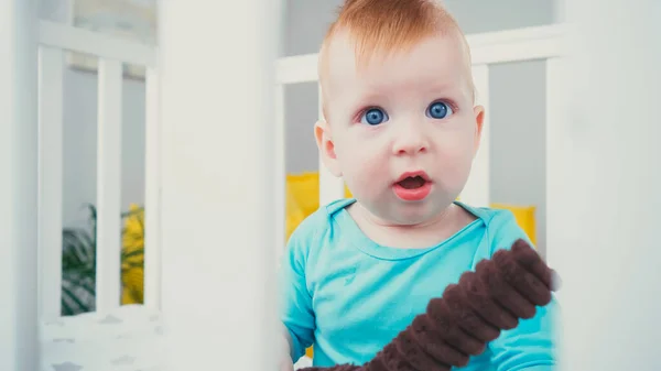 Surprised baby boy with blue eyes sitting in blurred baby crib with soft toy — Stock Photo