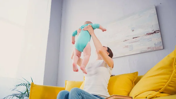 Low angle view of mother lifting barefoot infant son while sitting on sofa — Stock Photo