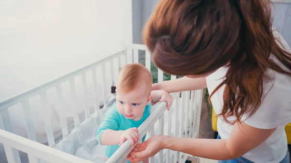 Morena mujer de pie cerca de bebé hijo en cuna - foto de stock