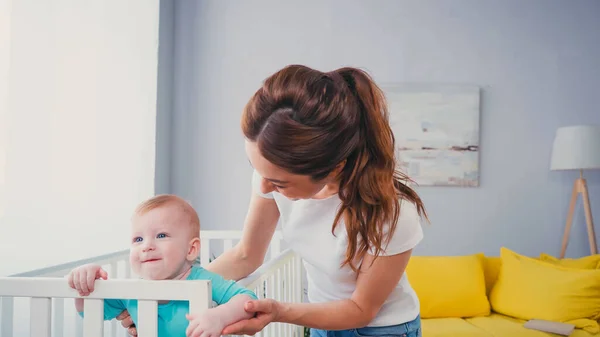 Brünette Frau steht neben lächelndem Säugling in Babybett — Stockfoto