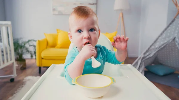 Bambino seduto in sedia di alimentazione e succhiare cucchiaio vicino ciotola — Foto stock