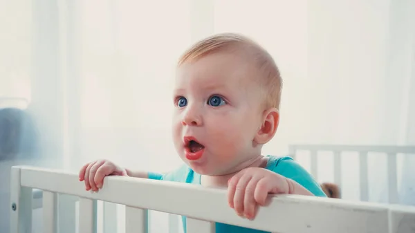 Surprised infant boy standing in baby crib and looking away — Stock Photo