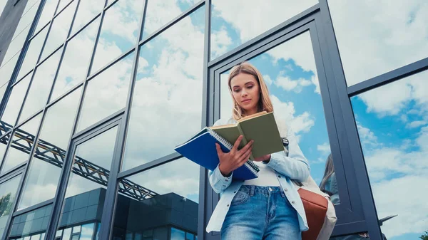 Low angle view of young student with notebooks and book standing near building on urban street — Stock Photo