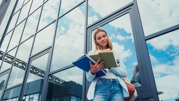 Tiefansicht einer glücklichen Frau mit Notizbüchern, die Buch liest und in der Nähe eines Gebäudes an der städtischen Straße steht — Stockfoto