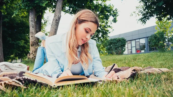 Joven estudiante escribiendo cerca de libro mientras está acostado en manta en el parque - foto de stock