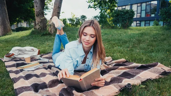 Alegre estudiante leyendo libro mientras que miente en manta a cuadros en parque - foto de stock