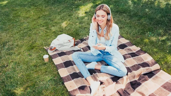 Vista de ángulo alto de estudiante feliz en auriculares con teléfono inteligente en manta en el parque - foto de stock