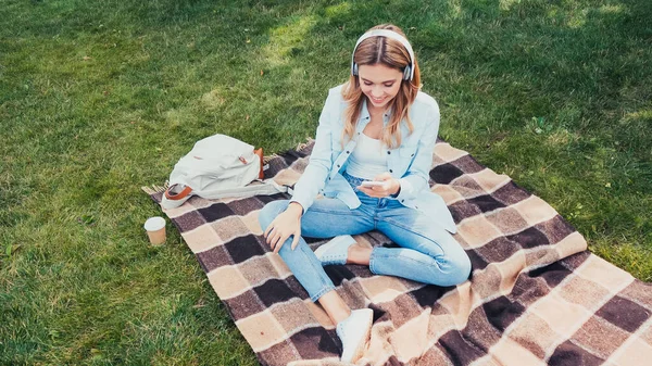 Visão de alto ângulo da mulher sorridente em fones de ouvido usando smartphone no cobertor no parque — Fotografia de Stock