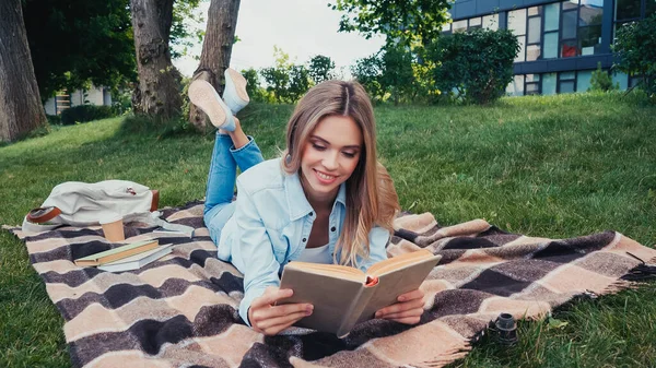 Jovem estudante alegre ler livro enquanto deitado no cobertor quadriculado no parque — Fotografia de Stock