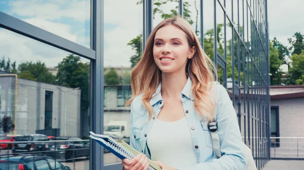 Pleased young student holding notebooks near building — Stock Photo