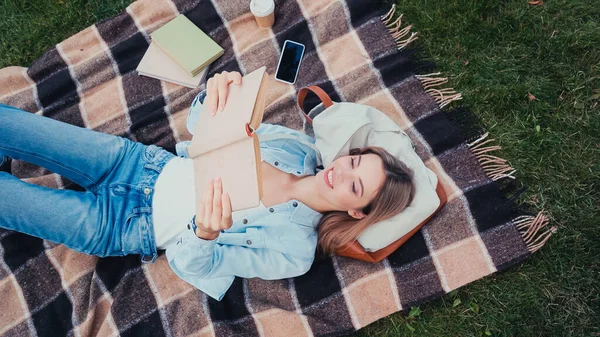 Вид сверху улыбающегося студента, читающего книгу и лежащего на одеяле в парке — стоковое фото