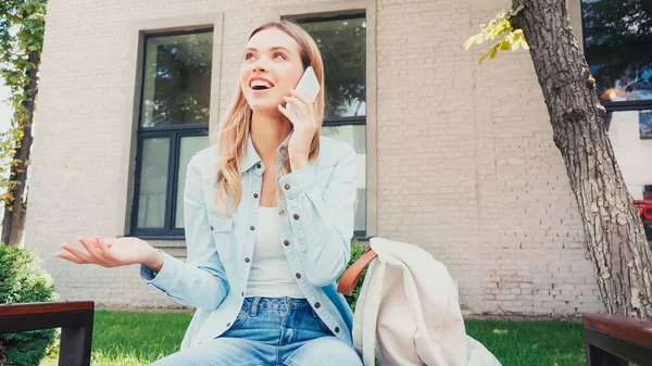 Smiling student talking on smartphone near backpack on bench — Stock Photo