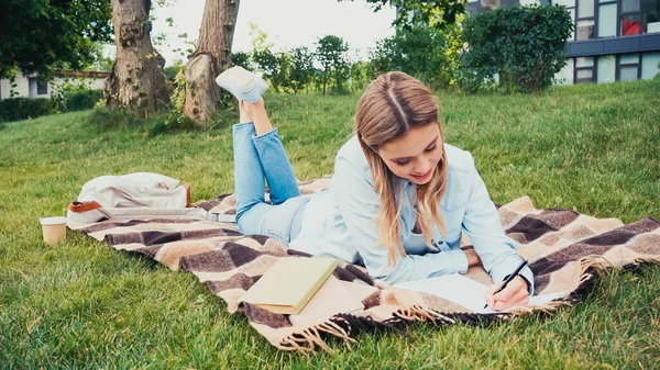 Fröhliche Studentin schreibt in Notizbuch neben Büchern, während sie auf Decke im Park liegt — Stockfoto