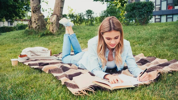 Feliz estudiante escribiendo en cuaderno y leyendo libro mientras está acostado en la manta en el parque - foto de stock