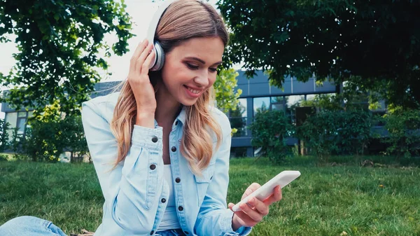 Donna felice utilizzando smartphone e ascoltare musica in cuffie wireless nel parco — Foto stock