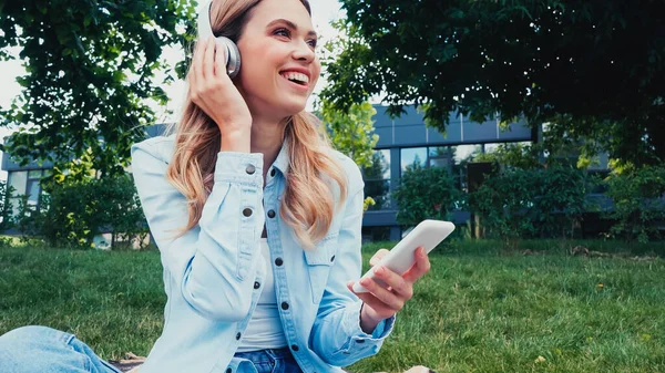 Cheerful woman using smartphone and listening music in wireless headphones in park — Stock Photo