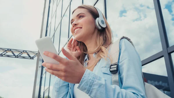 Low angle view of happy woman using smartphone and listening music in wireless headphones near building — Stock Photo