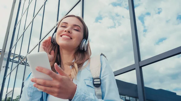 Vue à faible angle de la jeune femme gaie en utilisant un smartphone et écouter de la musique dans les écouteurs sans fil près du bâtiment — Photo de stock