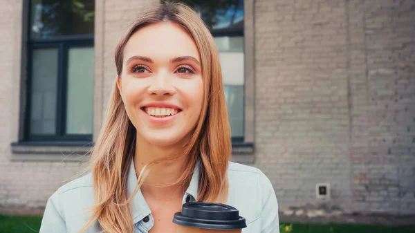 Positive woman holding coffee to go and smiling near building on urban street — Stock Photo