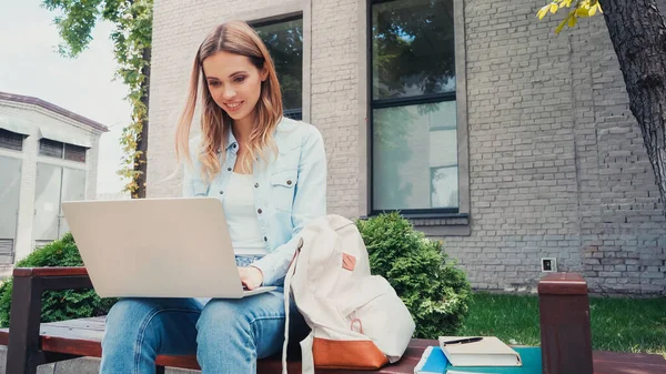 Studente sorridente utilizzando laptop vicino libro e quaderni su panchina — Foto stock