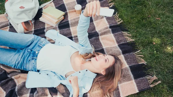 Vista superior del estudiante sacando la lengua mientras toma selfie cerca de libros en manta en el parque - foto de stock