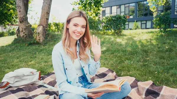 Feliz estudiante saludando la mano y sosteniendo libro en el parque - foto de stock