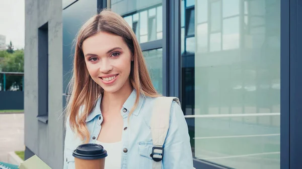 Happy woman holding coffee to go and smiling near building on urban street — Stock Photo