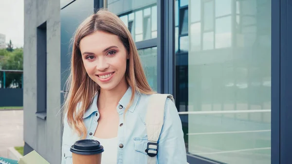 Mulher feliz segurando café para ir e sorrindo perto de construção na rua urbana — Fotografia de Stock