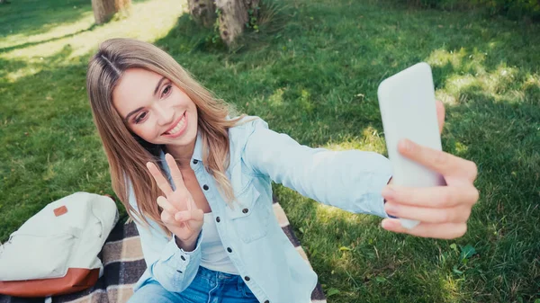 Estudiante sonriente mostrando señal de paz mientras toma selfie en el parque - foto de stock