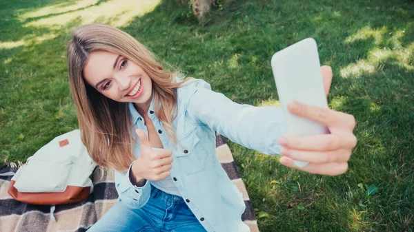 Estudiante feliz mostrando el pulgar hacia arriba mientras toma selfie en el parque - foto de stock