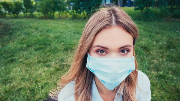 Junge Frau mit blauer medizinischer Maske blickt im grünen Park in die Kamera — Stockfoto